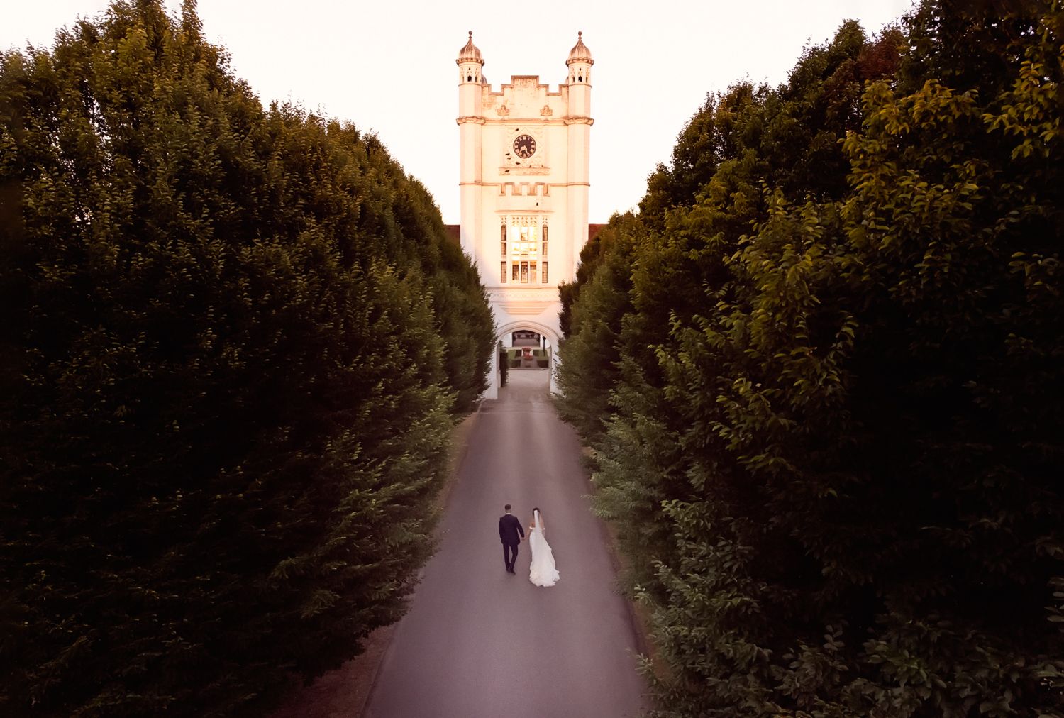 A couple walks down a tree-lined path leading to a majestic church tower in a romantic wedding photo.