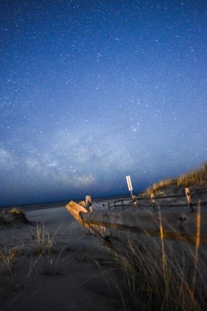 Starlit night sky over beach dunes with wooden posts illuminated by warm light.