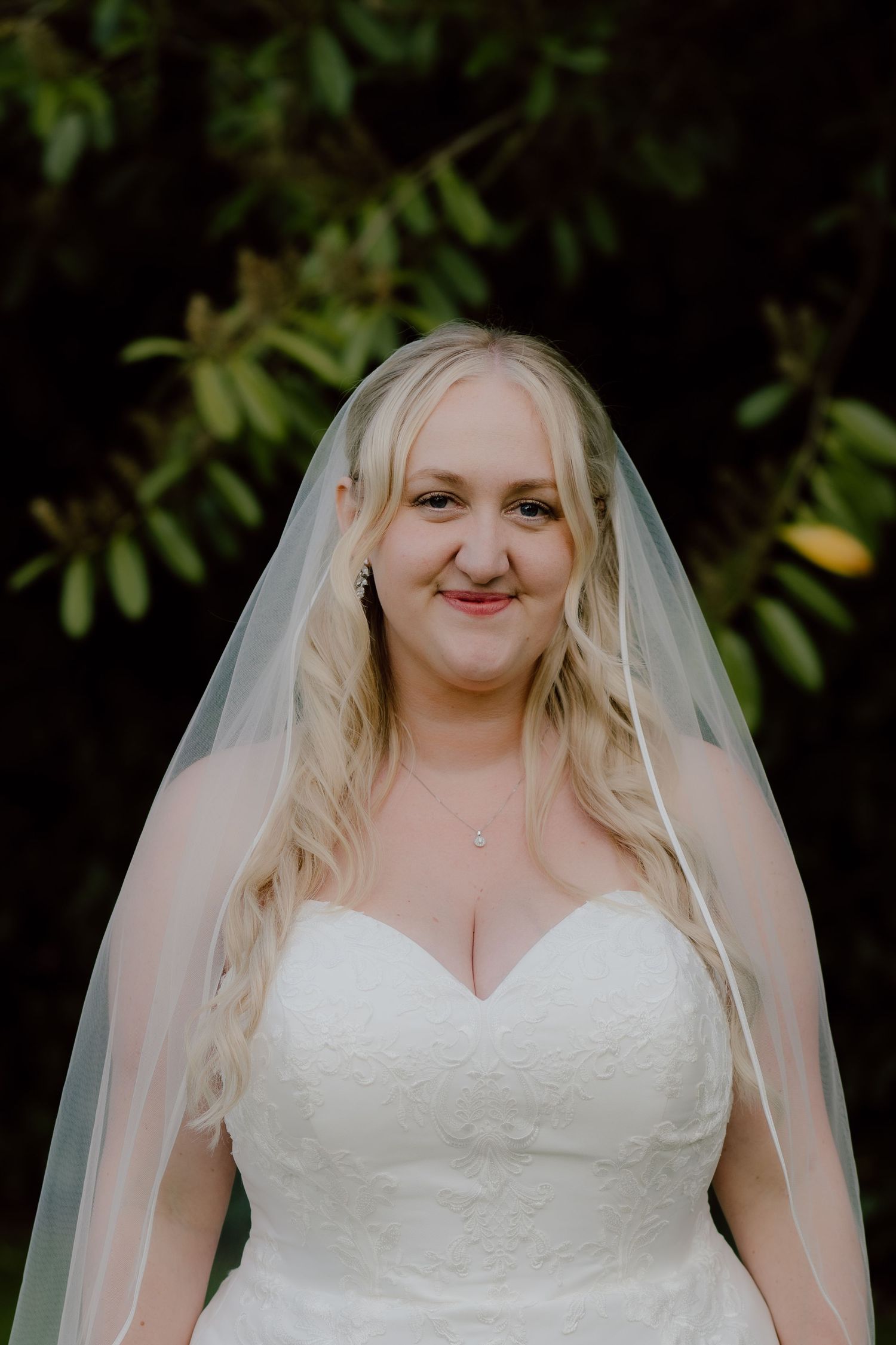 A bride in a strapless wedding gown and veil smiles against a dark foliage background.