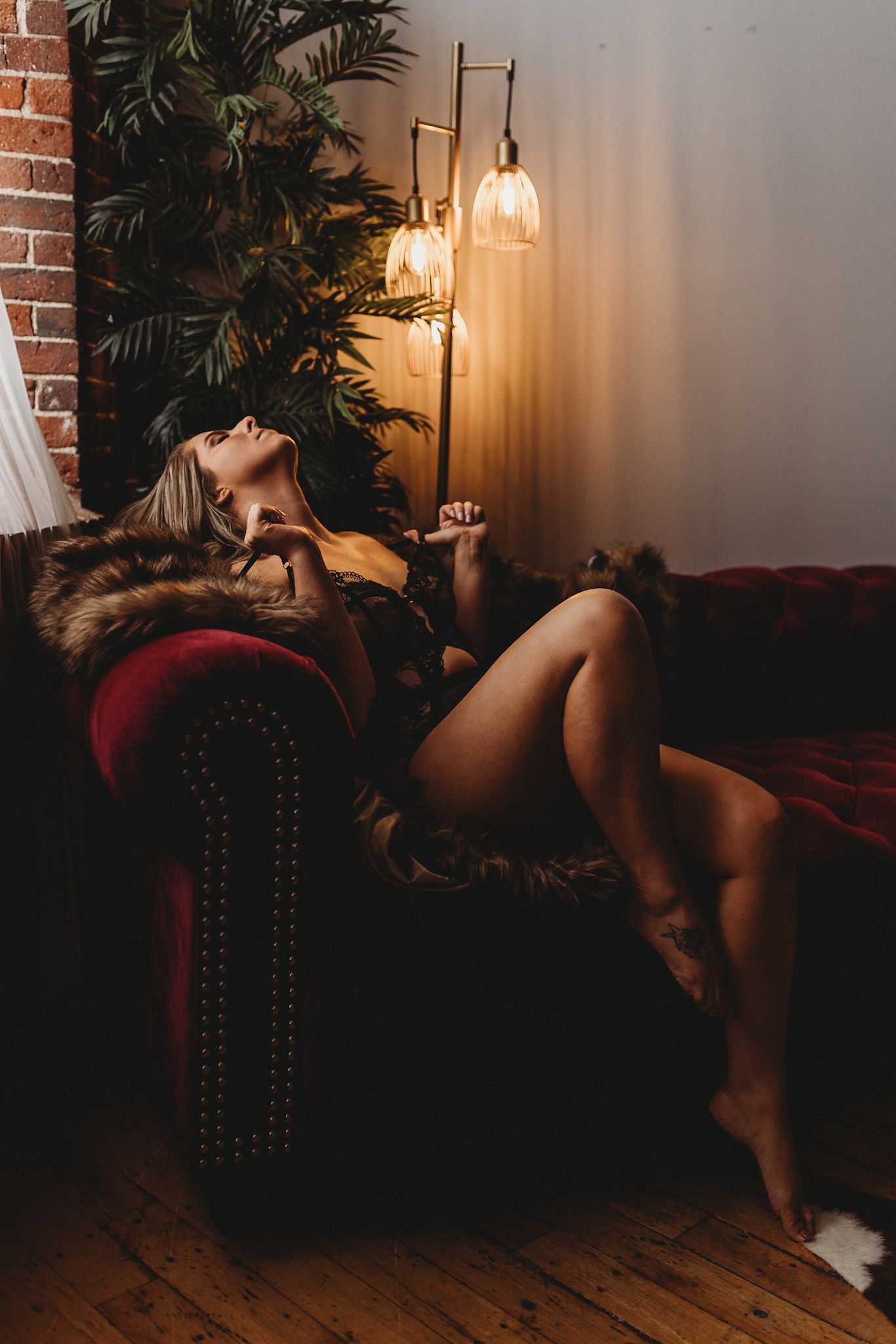 Artistic boudoir photo on red velvet couch with mood lighting and plants.