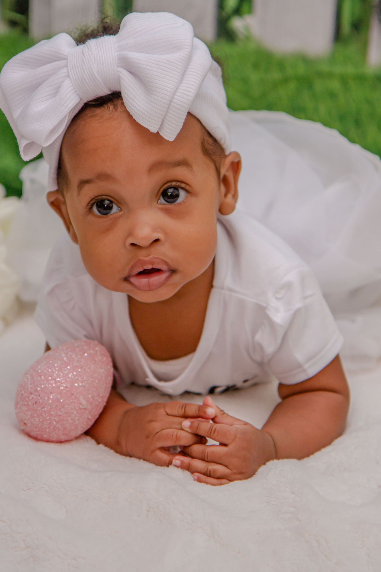 Baby wearing white outfit with bow headband lies on white blanket.
