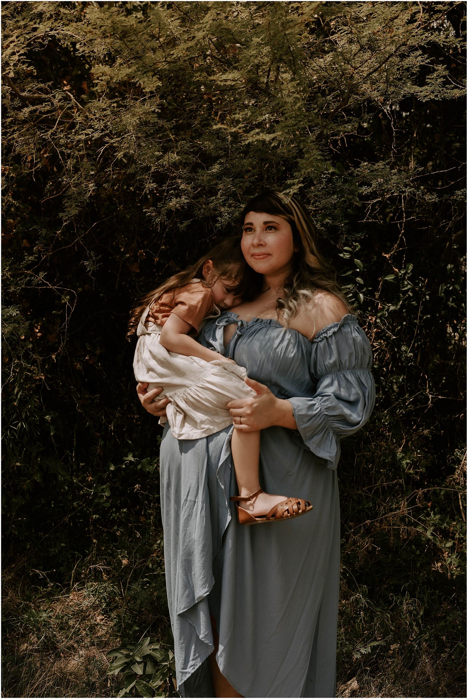 Mother in flowing blue dress holds child in peach outfit against dark forest background.