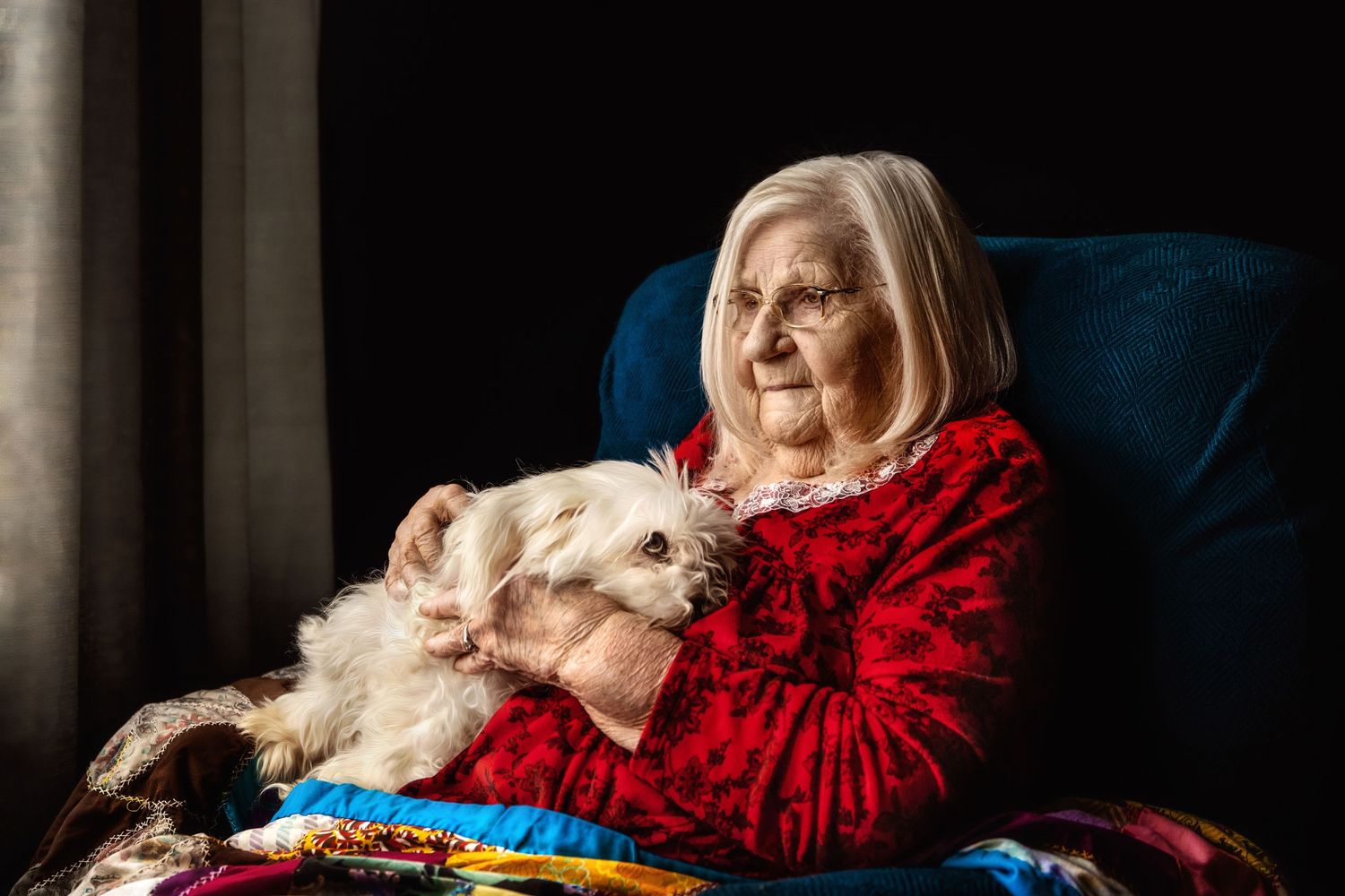 An elderly woman in a red sweater sits with a white dog in a blue armchair.
