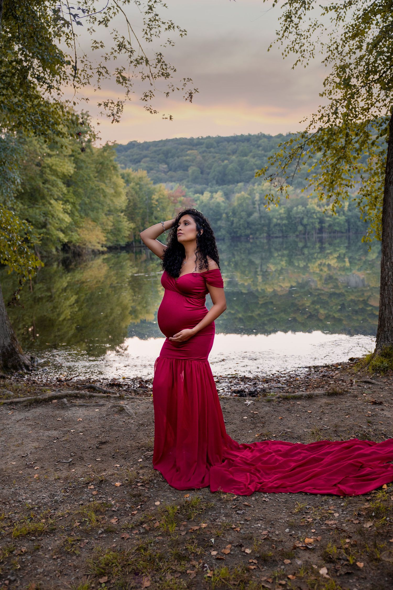 Pregnant woman in flowing red dress poses dramatically by lake at sunset.