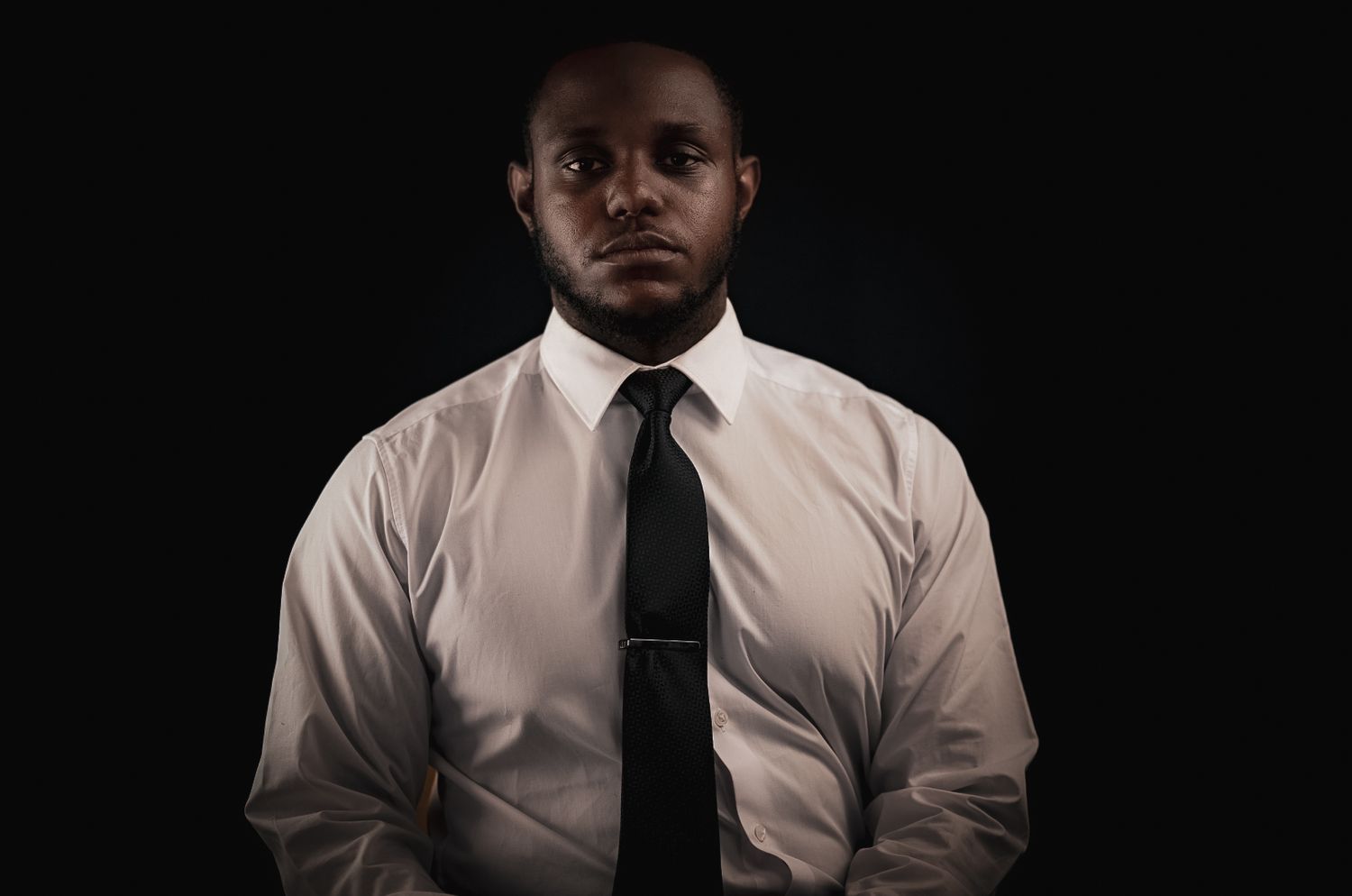 Professional headshot in white dress shirt and black tie against dark background.