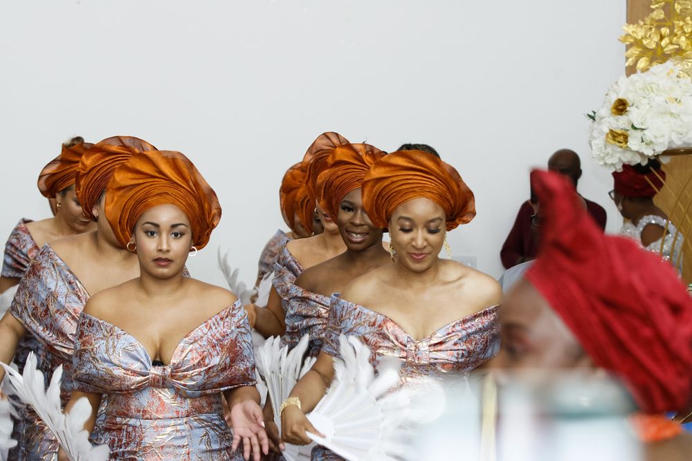 Attendees in traditional African dress with orange headwraps and off-shoulder gowns gather at a celebration.