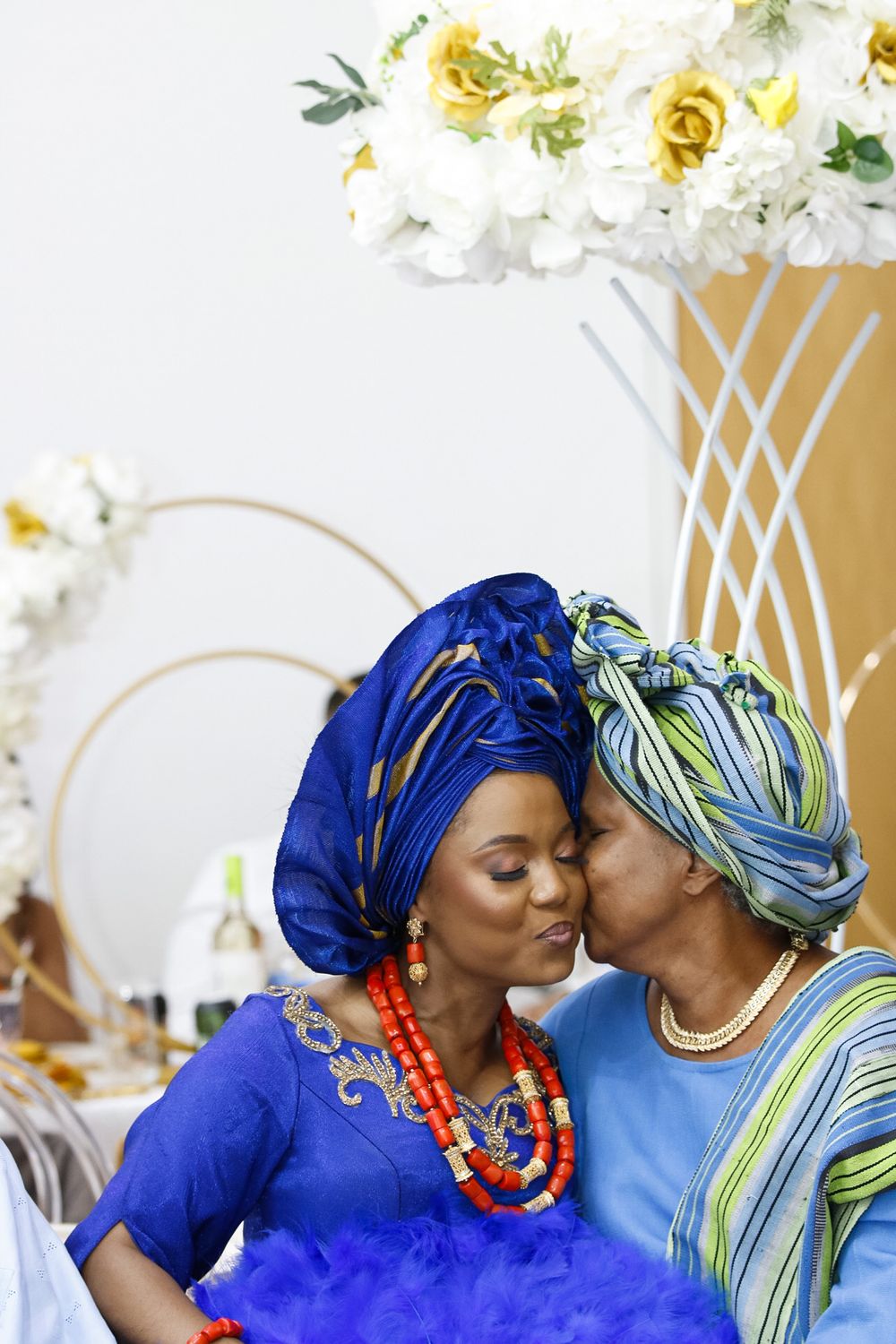 Two people in blue and patterned headwraps share a tender moment against a floral backdrop.