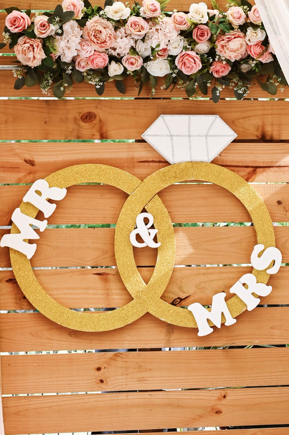 Gold interlocking rings with 'MR & MRS' text and pink roses on wooden backdrop make an elegant wedding decoration.