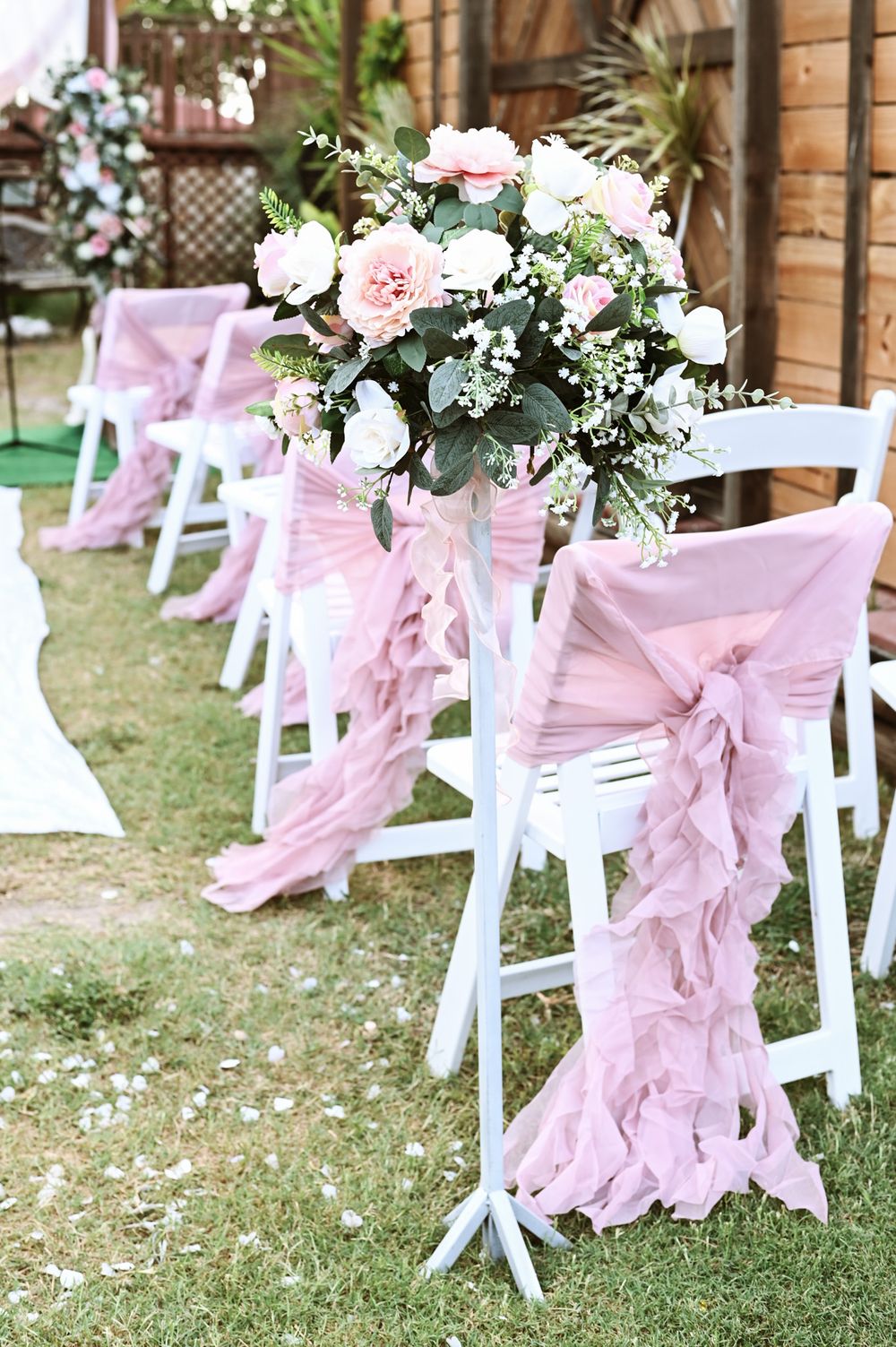 White folding chairs decorated with pink fabric sashes and floral arrangements for outdoor wedding ceremony.
