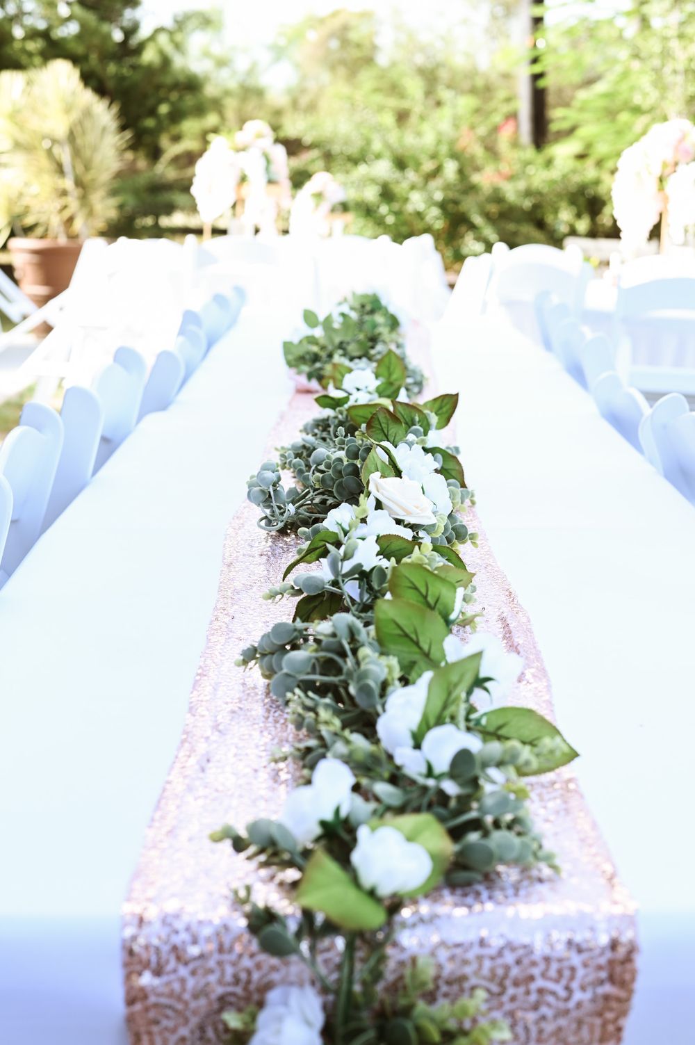 Long white reception table with floral garland runner and sequined decor.