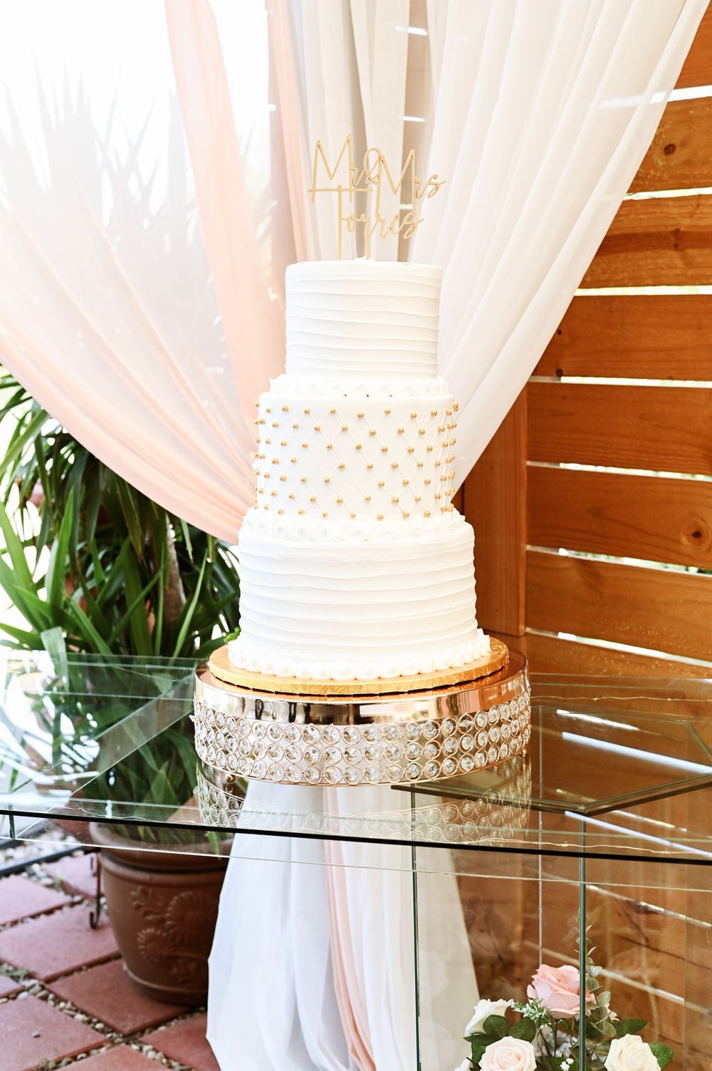 Elegant three-tier white wedding cake with pearl details on crystal stand and sheer drapery background.