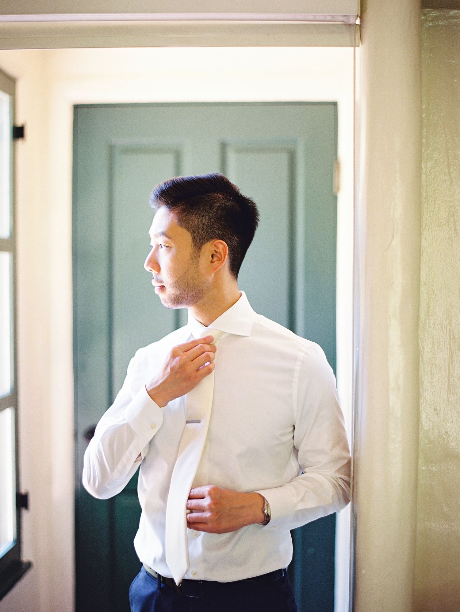 Wedding groom adjusting white dress shirt collar while getting ready in front of door. Allied Art Guild Wedding.