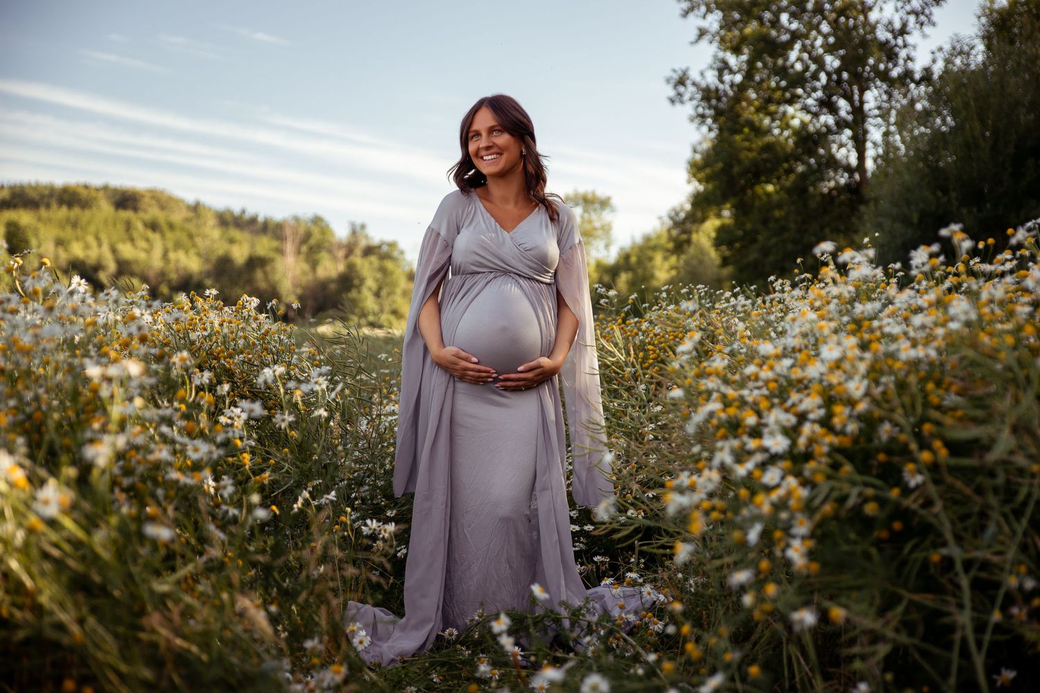 Expectant mother in flowing lavender dress poses in wildflower meadow at sunset.