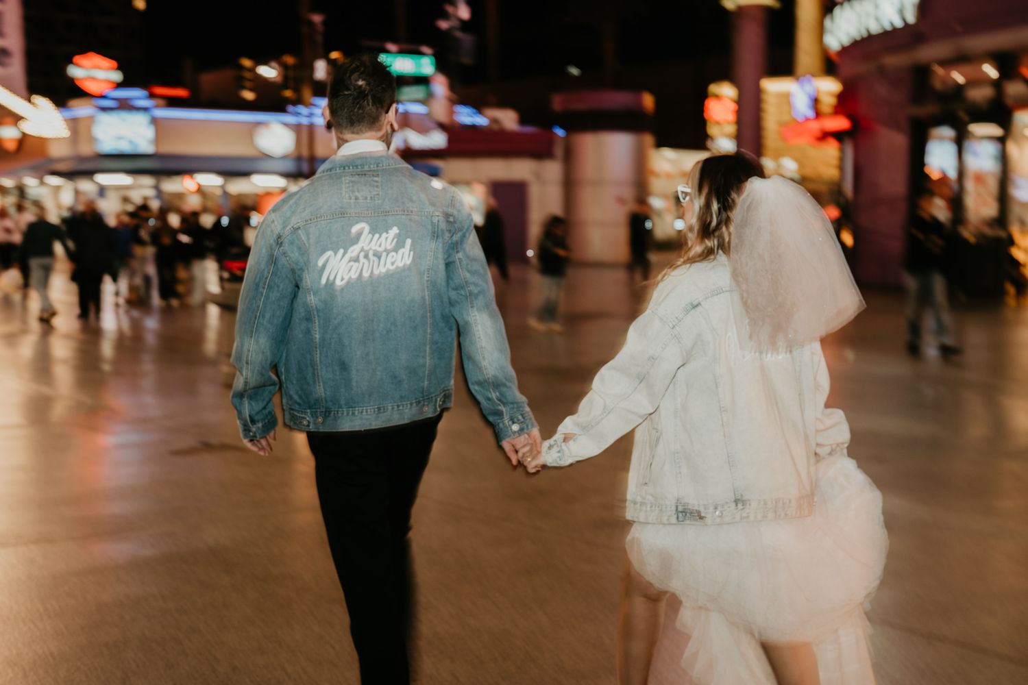 Wedding couple holding hands walking through casino in denim jacket and white dress.