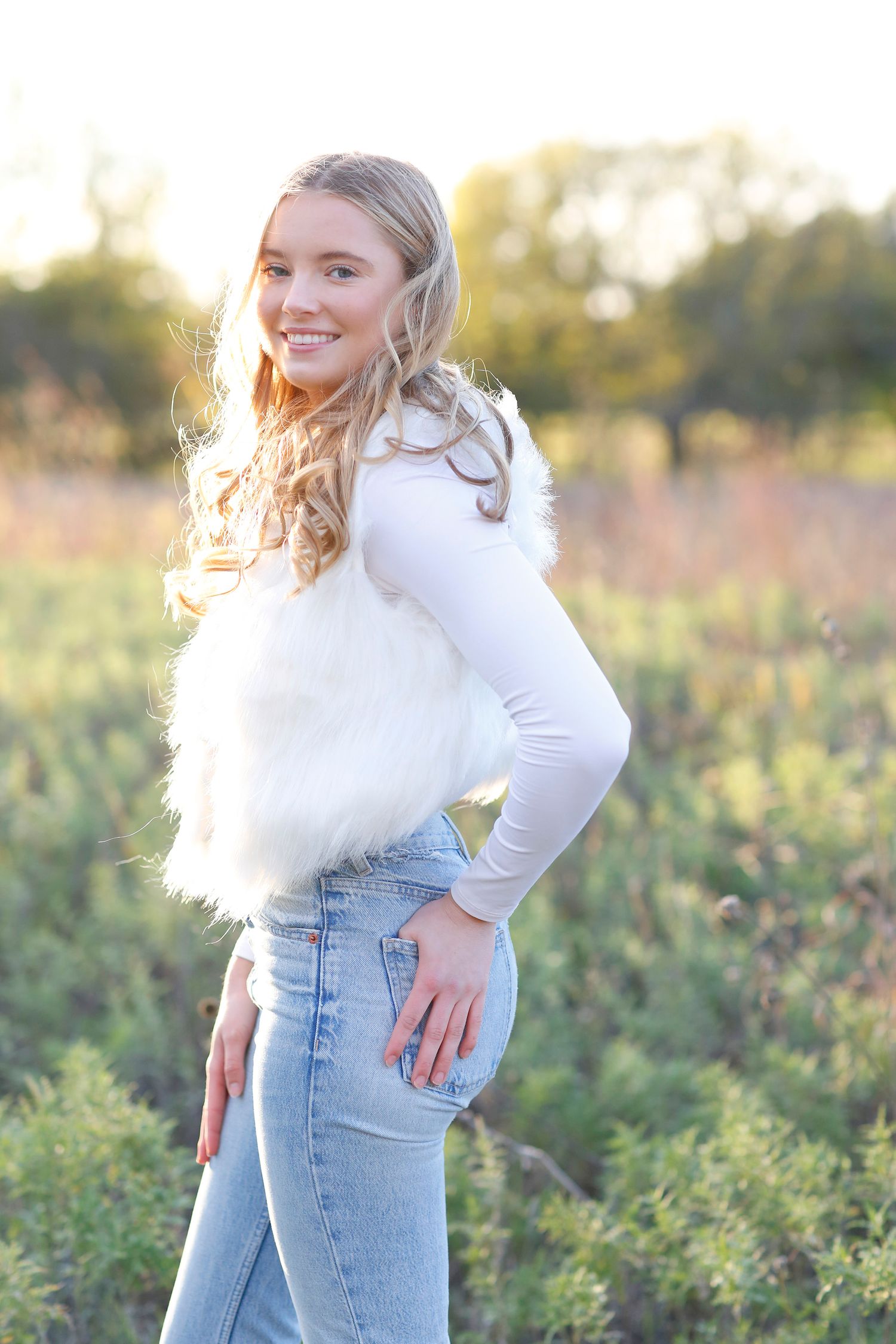 A person in a white fuzzy vest and jeans poses in a sunlit field at golden hour.