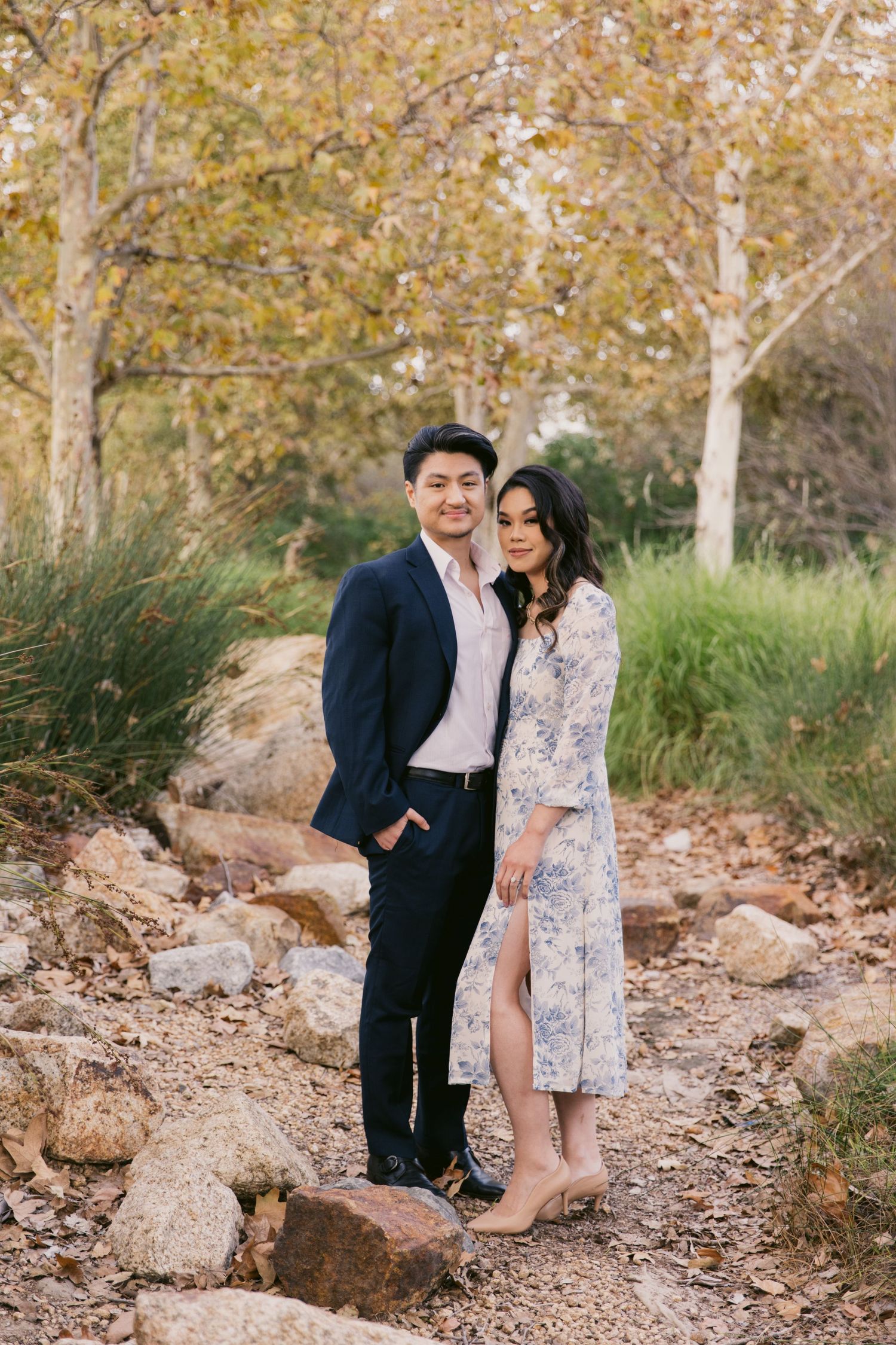 A couple poses together in a romantic outdoor setting with autumn trees and rocks in the background.