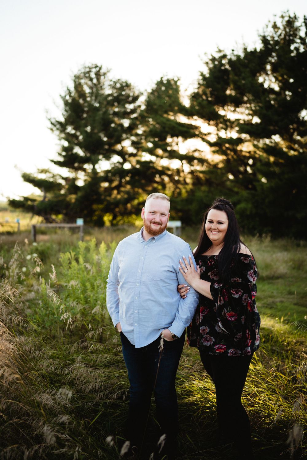 Couple standing together at sunset in a field with golden light filtering through pine trees.