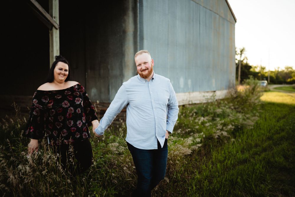A couple laughing together outside a metal barn during a countryside photoshoot.