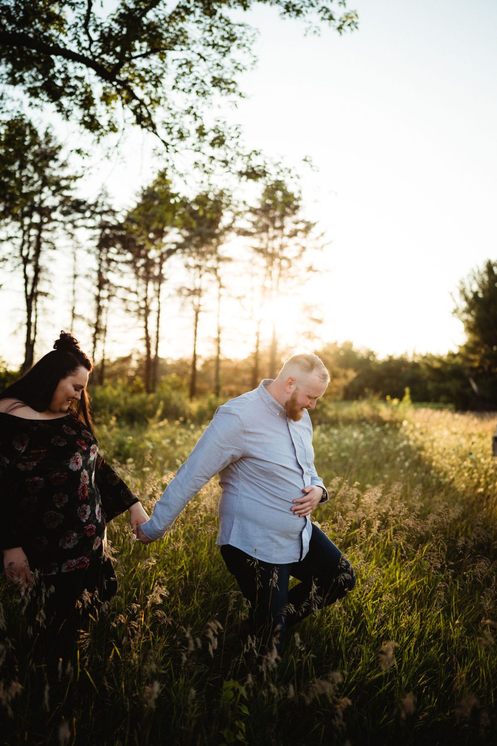 A couple walking through tall grass during golden hour with trees in background.