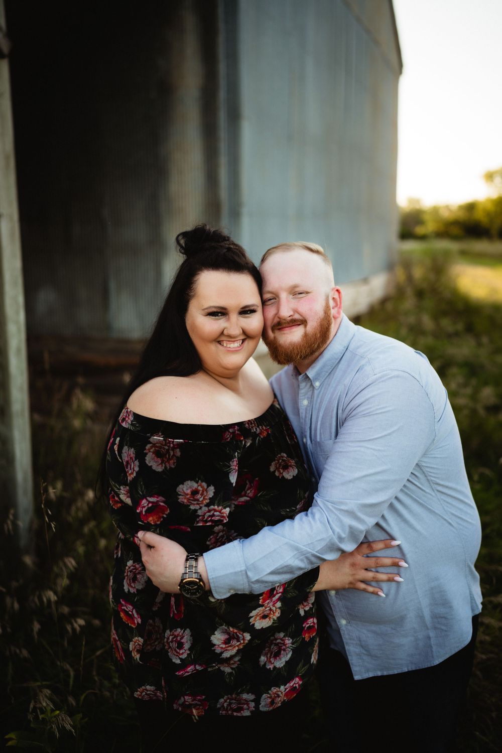 A couple embracing and smiling by a rustic metal barn during golden hour.