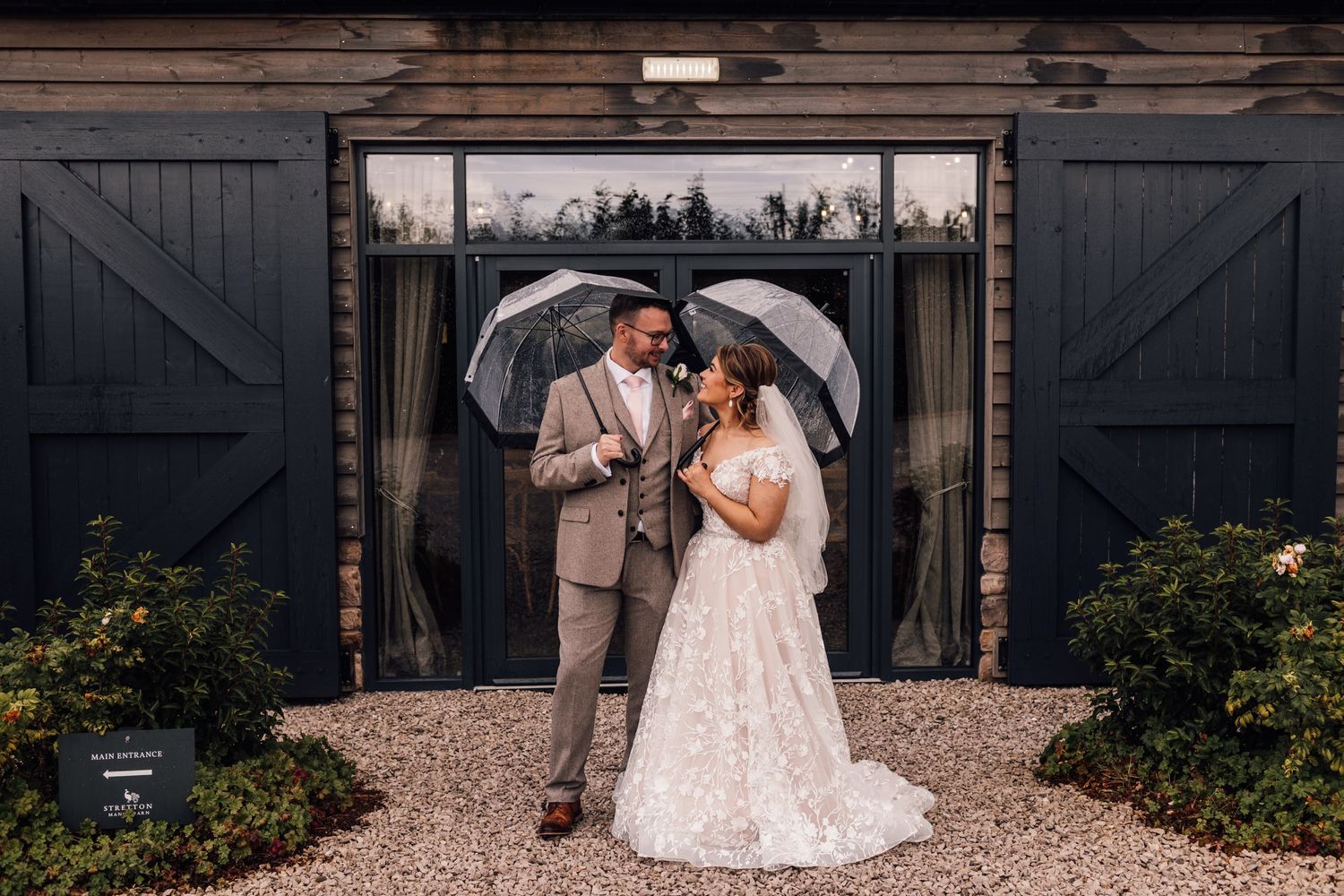 Newlyweds pose with umbrellas in front of barn doors surrounded by landscaping on gravel pathway.