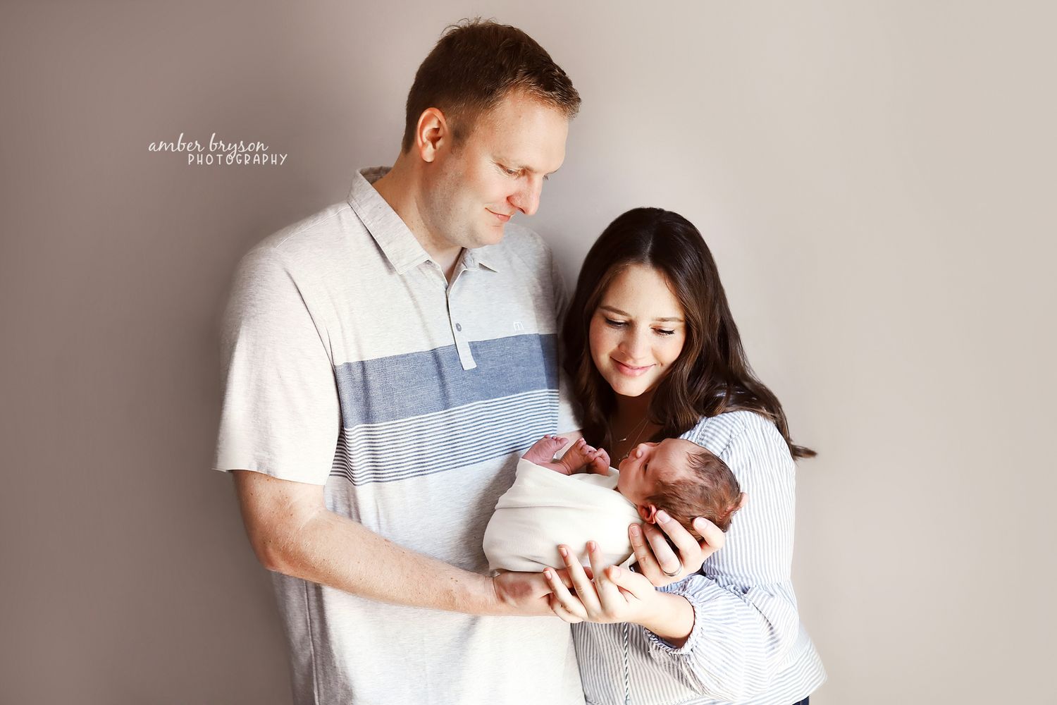 New parents holding their swaddled newborn against neutral wall background.