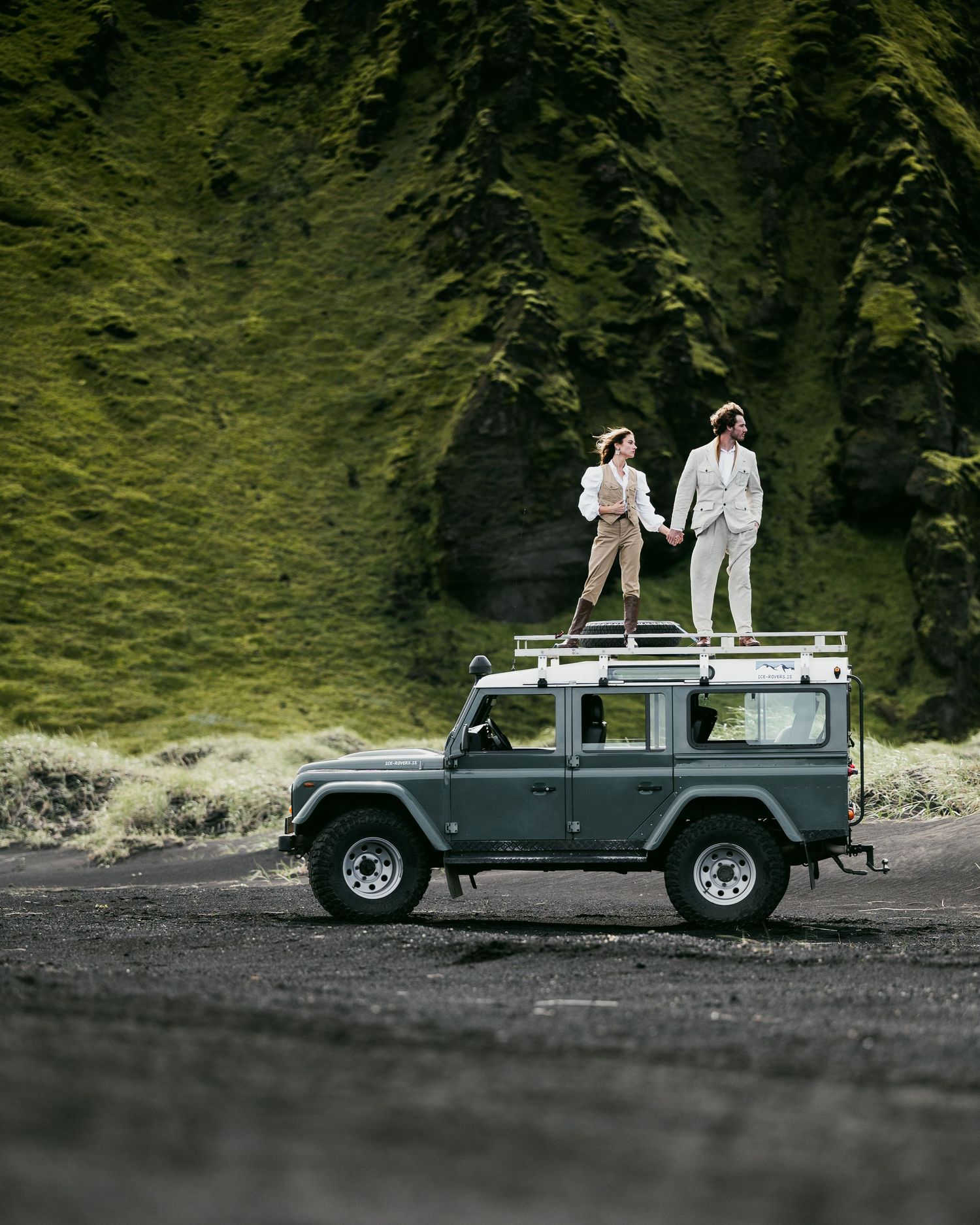A vintage Land Rover parked on a black sand beach with green cliffs in the background.