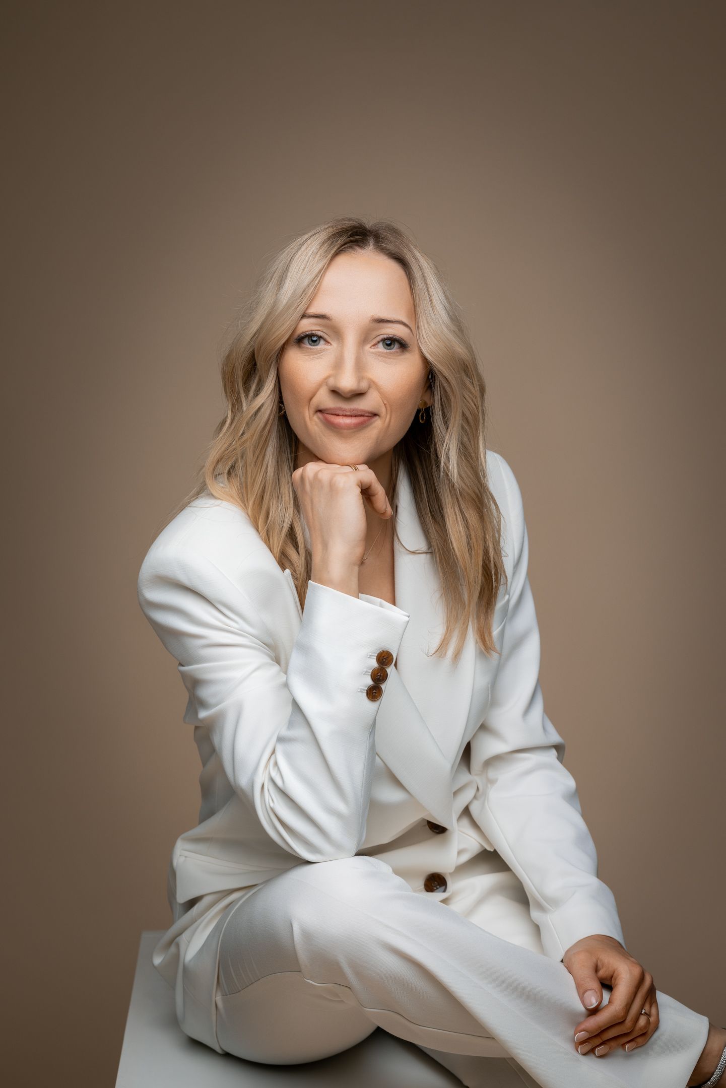Professional portrait of a person in a stylish white suit sitting casually against a brown background.