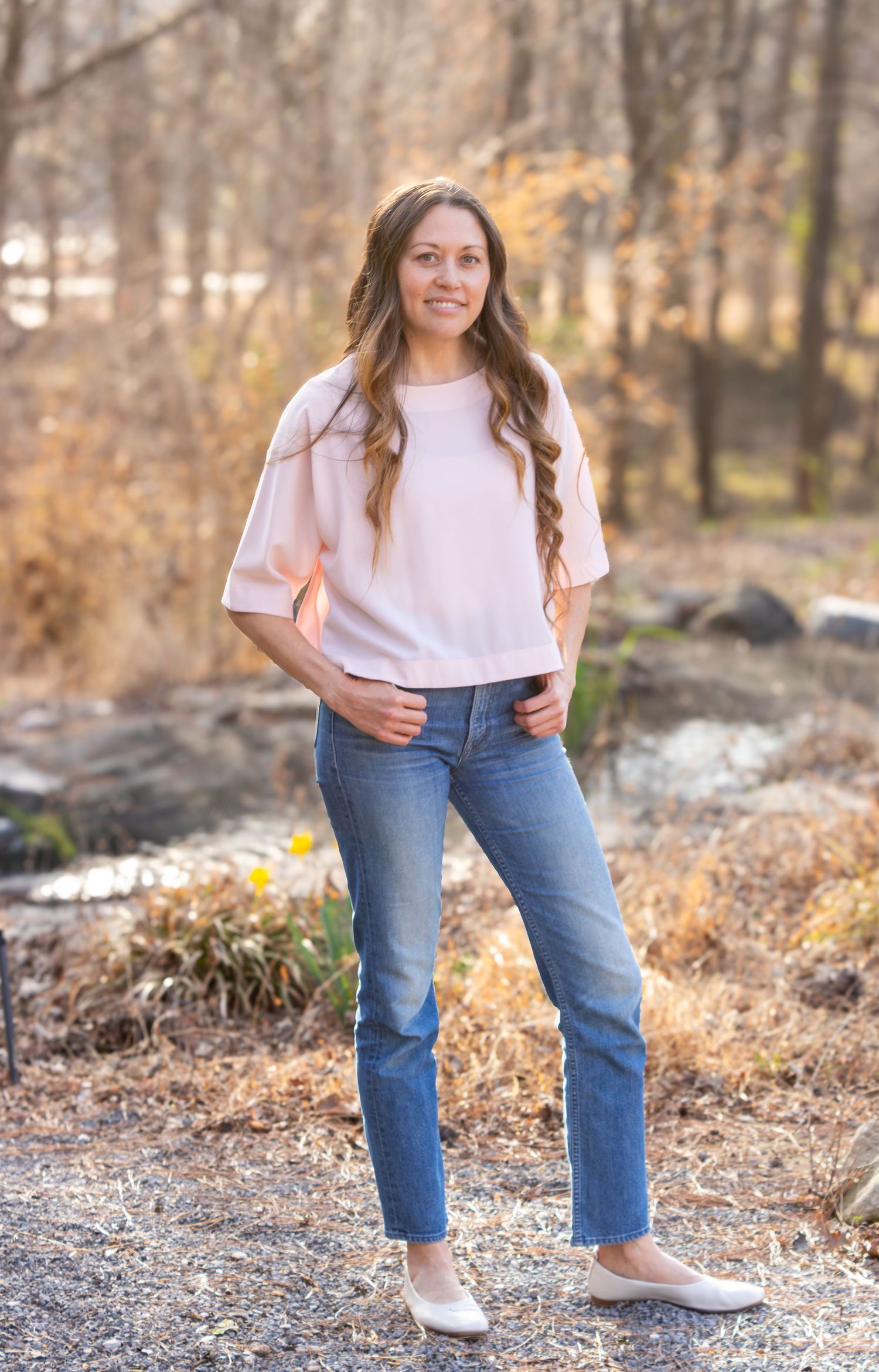 Girl in pink shirt and jeans poses outdoors in a scenic autumn forest setting.