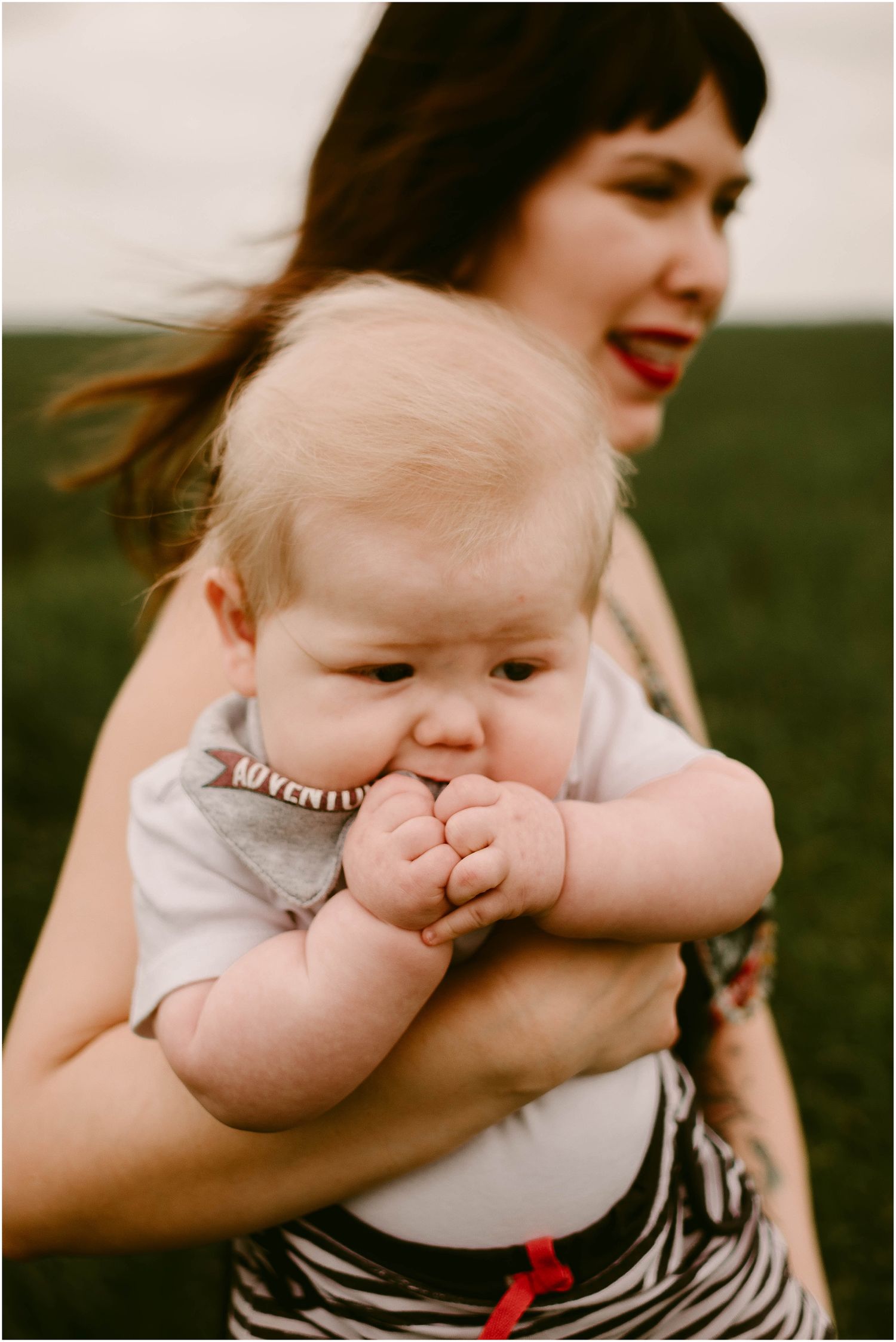 Baby with chubby cheeks puts fingers in mouth while being held outdoors on a cloudy day.