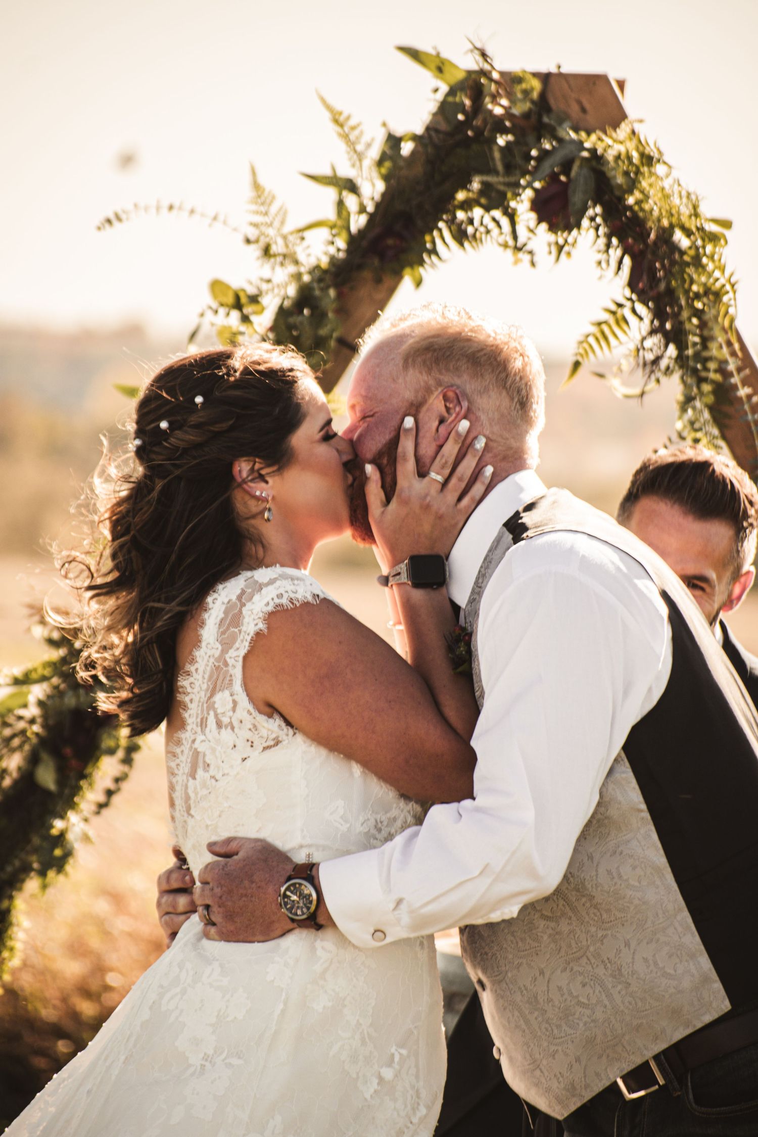 Romantic couple embraces under floral arch during sunset wedding ceremony.