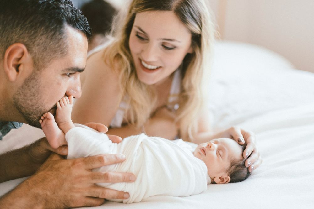 Intimate family moment with baby on white bedding in warm natural light.