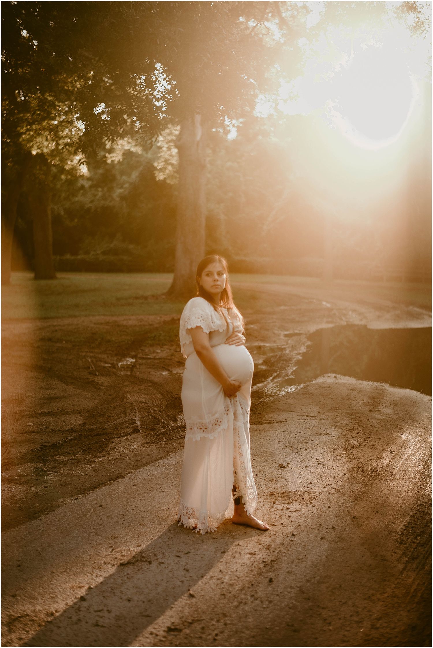 Golden hour maternity portrait on rustic path with sunlit trees.