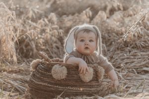 Baby in bunny costume peeks over edge of woven basket in rustic outdoor setting.