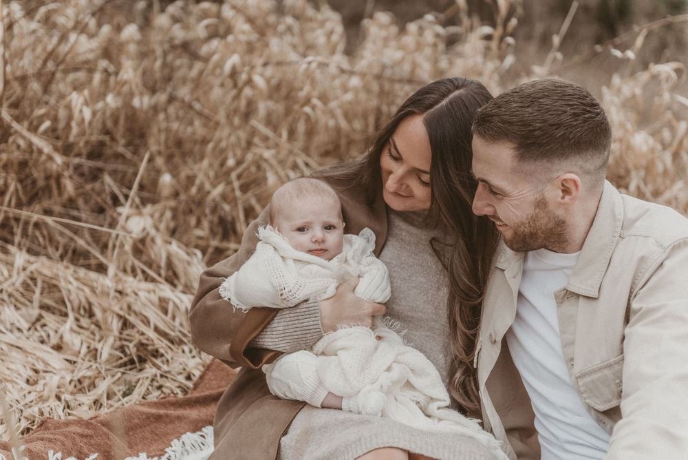 A family sitting in tall grass sharing joyful moments with their baby in neutral tones.