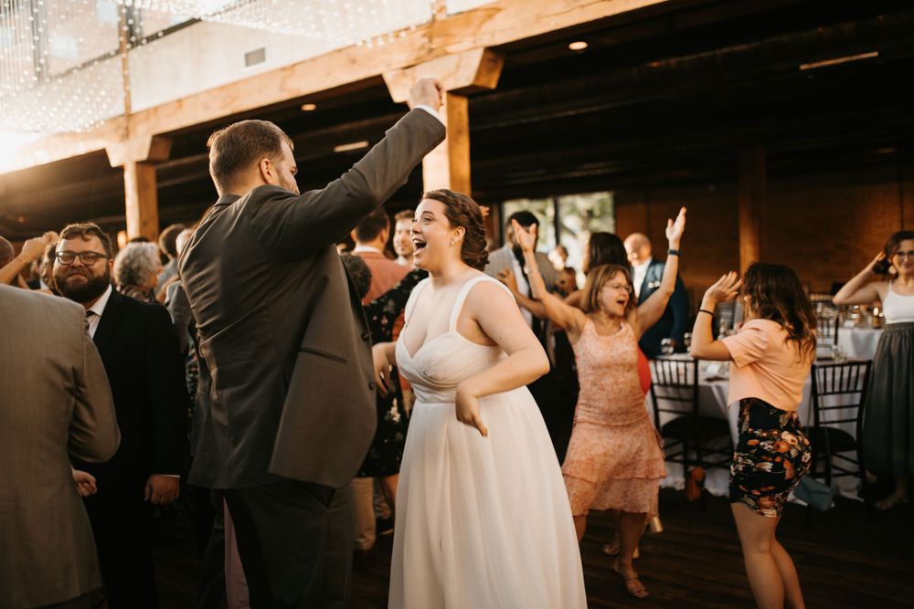 Bride in white gown and groom dance energetically with cheering wedding guests at indoor reception.