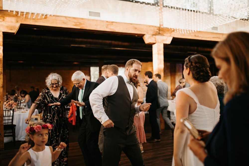 Wedding guests dance together at a rustic indoor reception venue with warm ambient lighting.