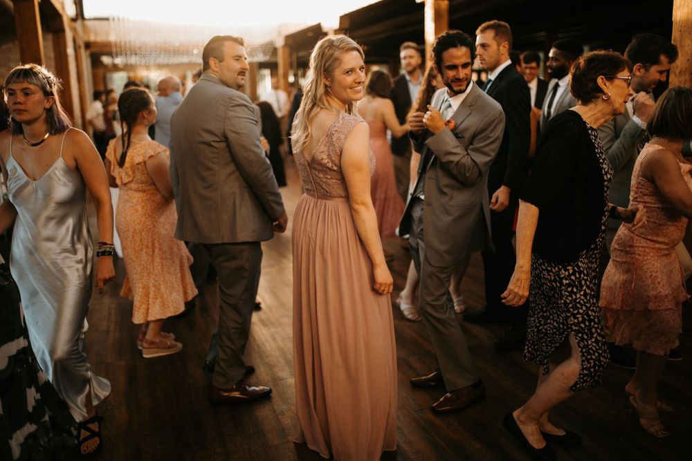 Smiling bridesmaid in dusty rose gown dances with guests at a warmly lit wedding reception.