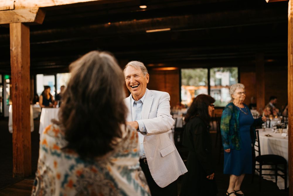 Older man in white blazer laughs joyfully while dancing with a woman at a wedding reception.
