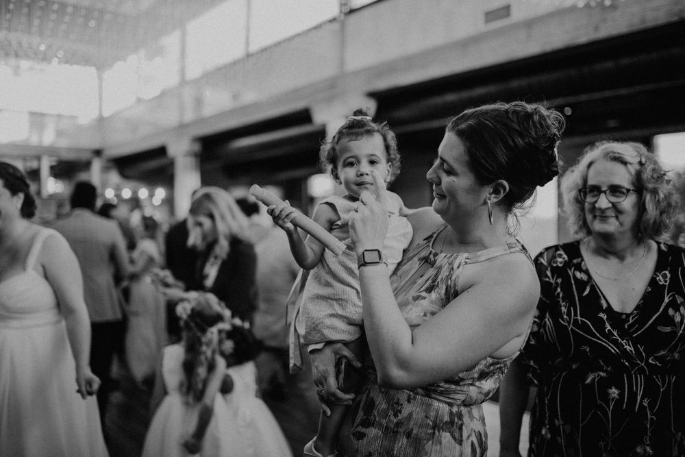 Woman holds a young toddler girl while standing among wedding guests in black and white photo.
