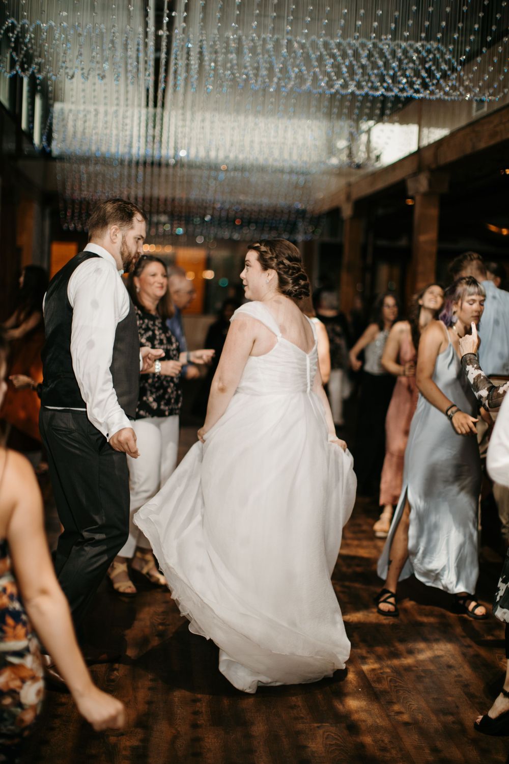Bride twirls in white gown with groom on crowded dance floor under crystal chandelier at reception.