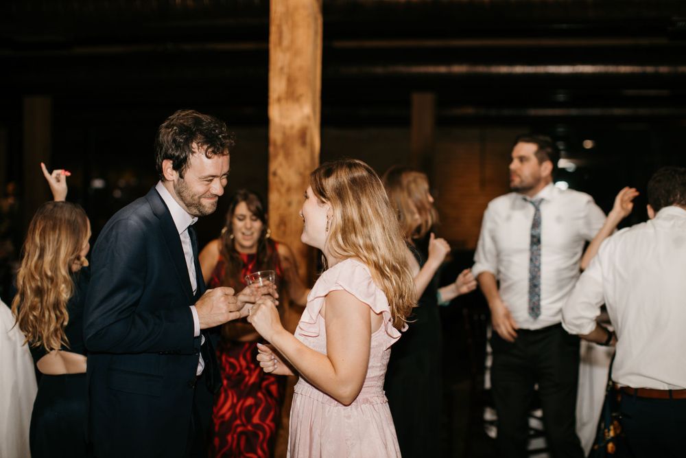 Couple dances closely together smiling at a lively wedding reception with guests in background.