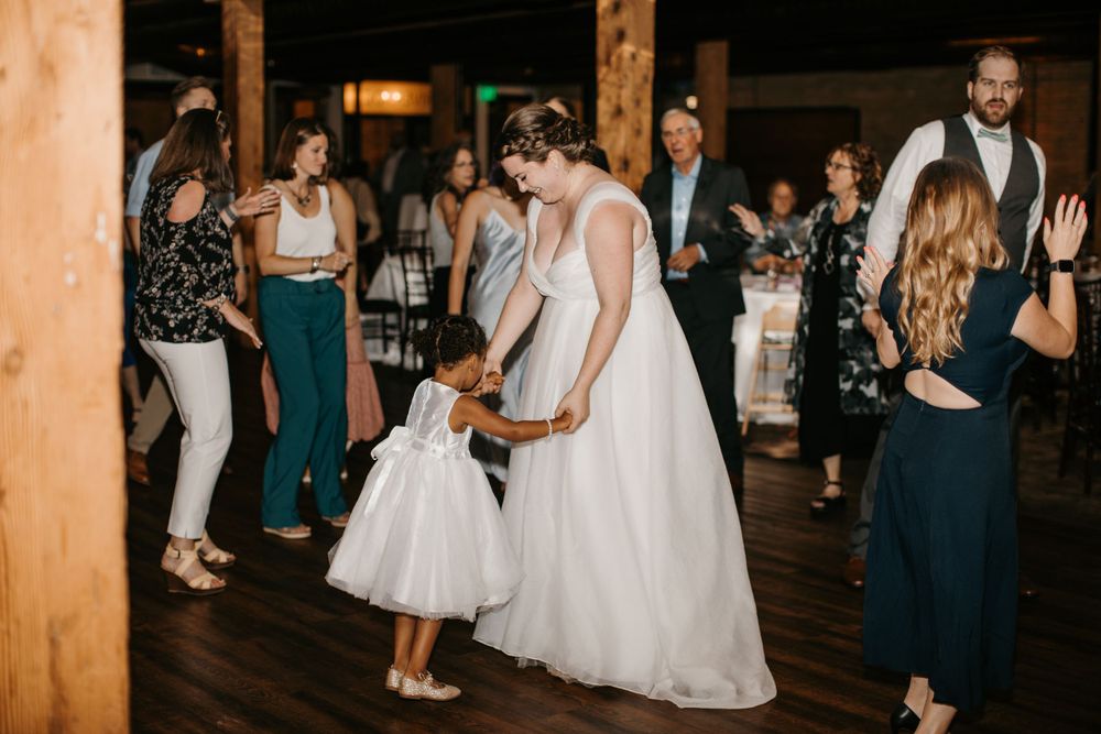 Bride in white gown dancing with flower girl in white dress at rustic wedding reception.