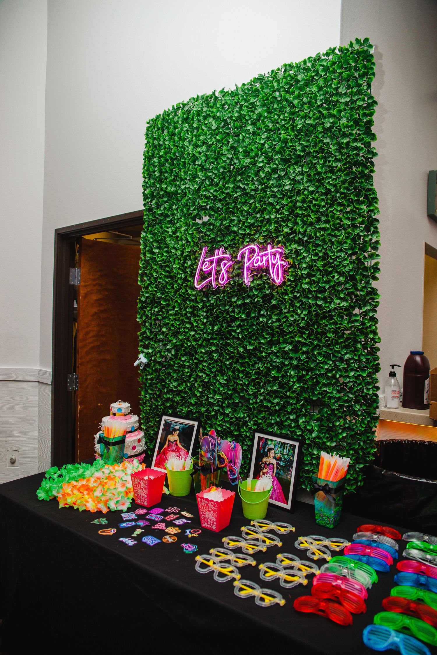 Photo backdrop with green leaf wall and colorful party decorations on black table.