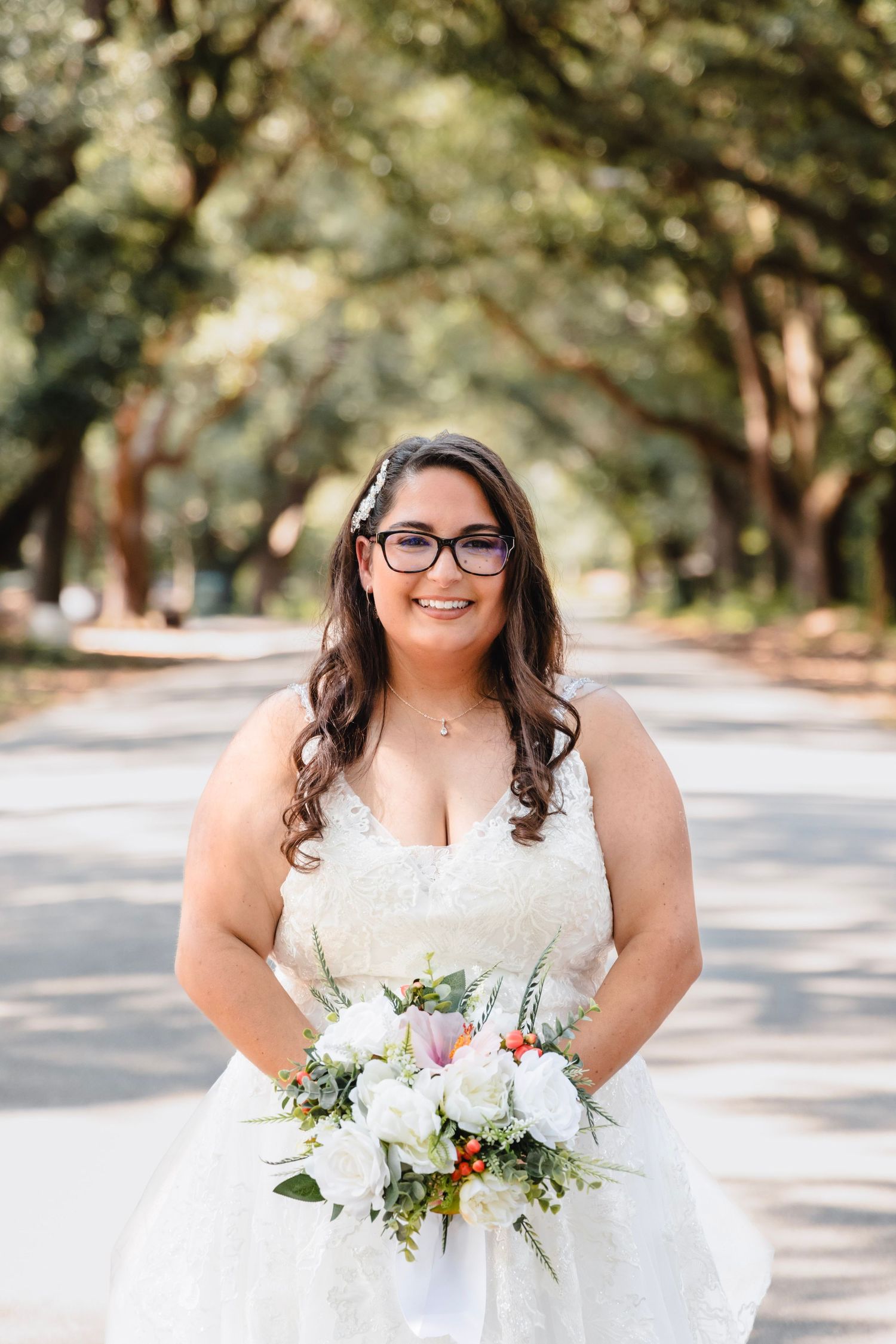 A bride in a white wedding dress and glasses holds a beautiful bouquet of white and pink flowers on a tree-lined path.