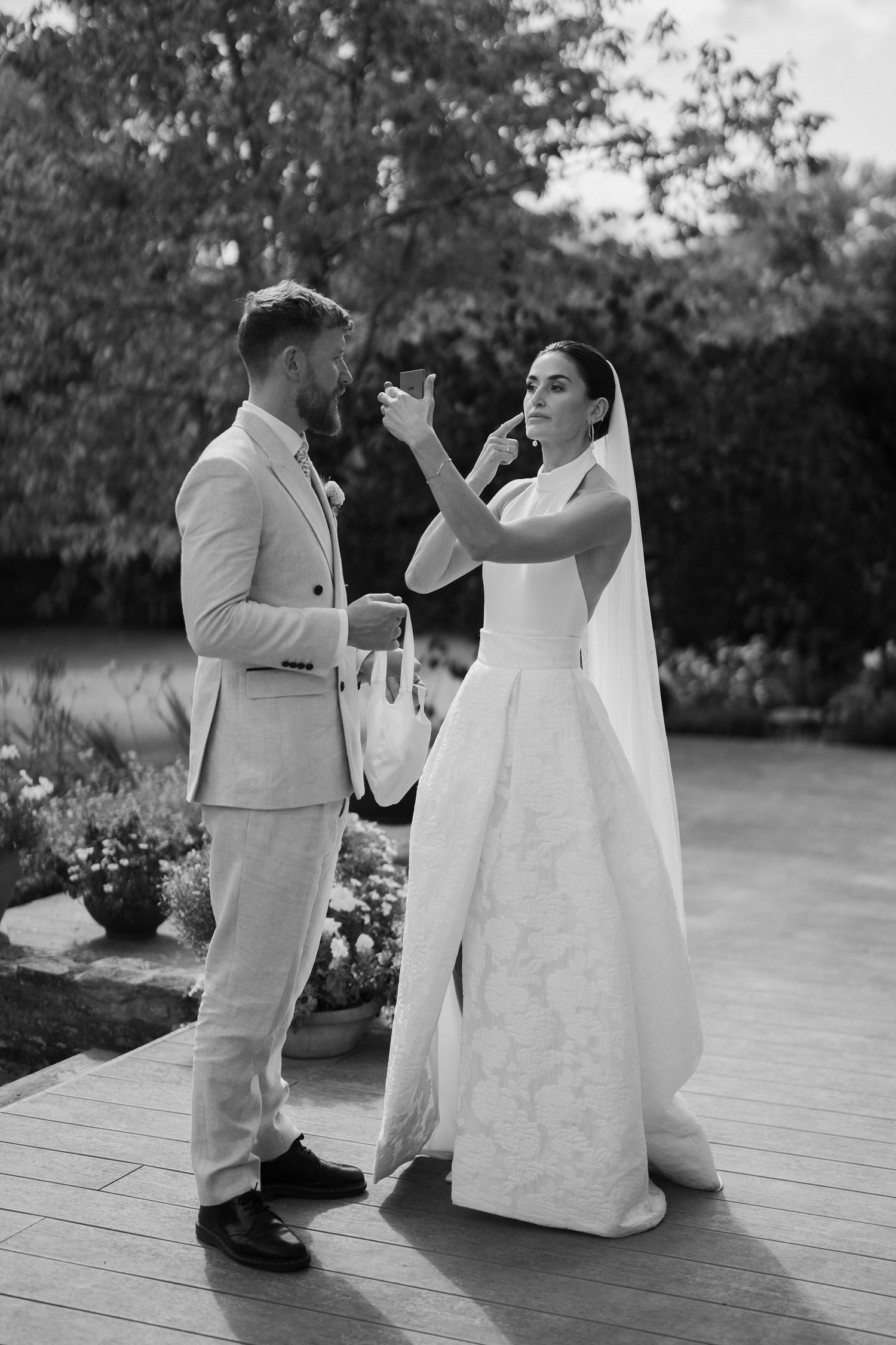 bride adjusting maker up in mirror while groom holds her bag in gardens in islip, oxfordshire