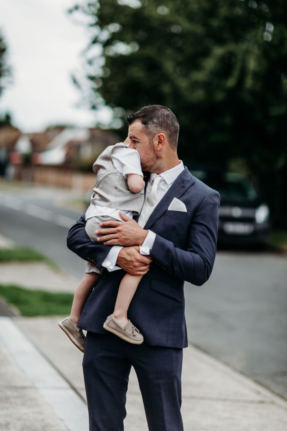 Person in navy suit holds small child wearing grey outfit on suburban street.