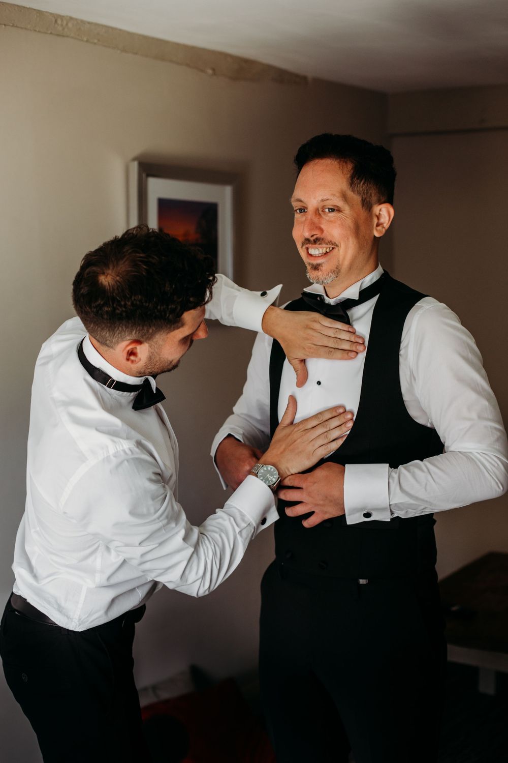 Two people in formal attire laugh and help each other with their wedding day preparations.