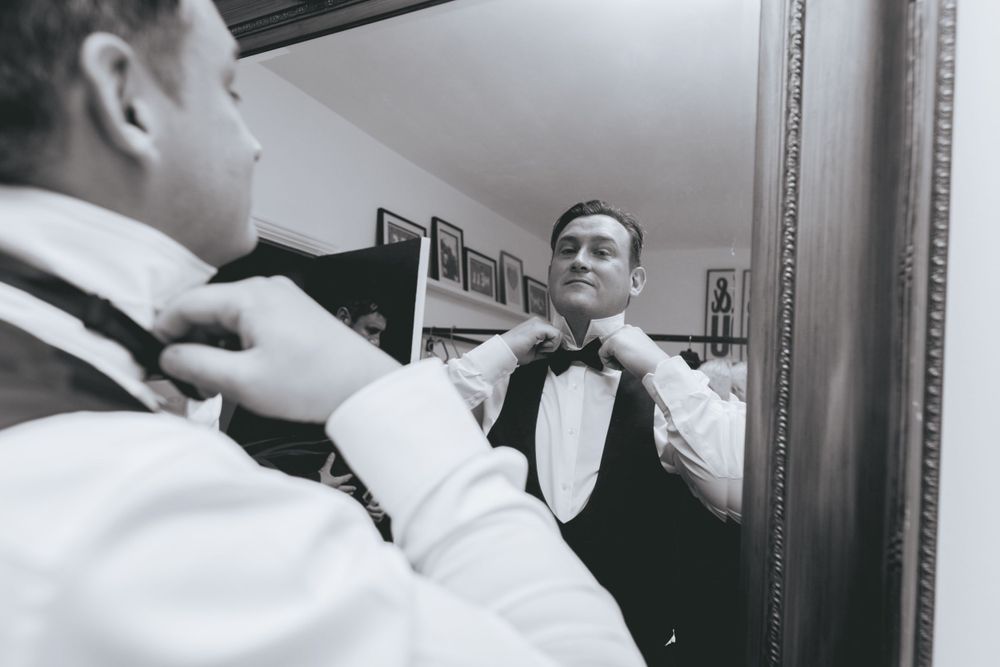 Black and white photo of man adjusting bow tie in mirror while getting ready.