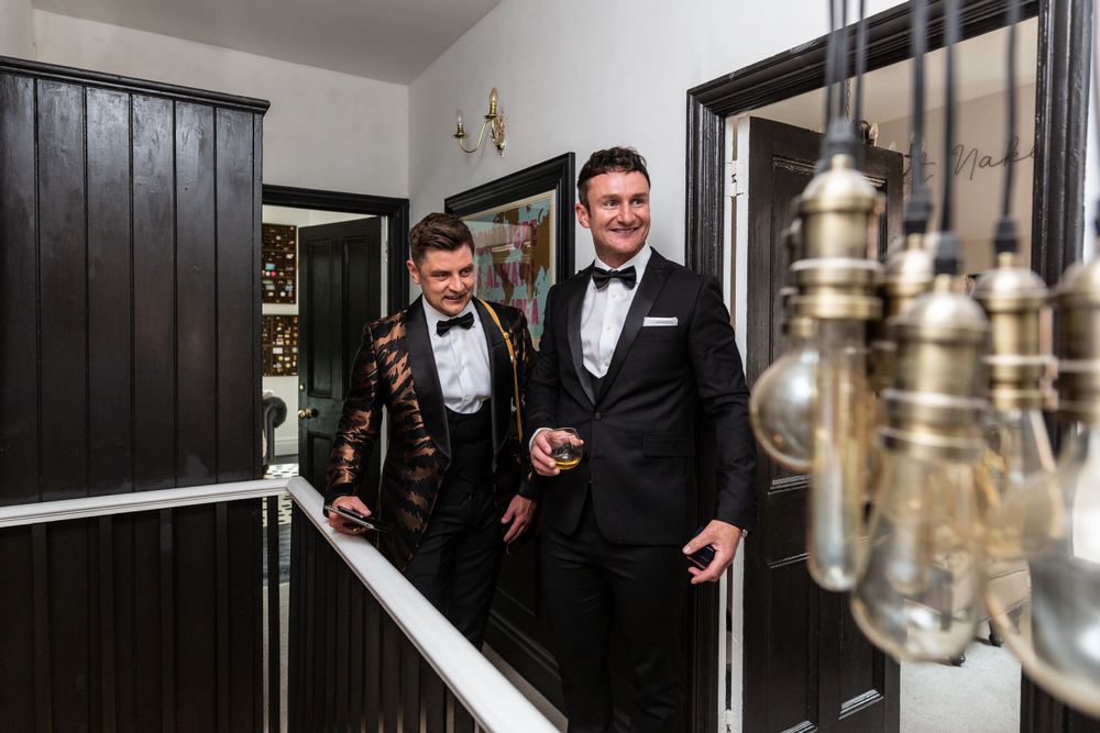 Two people in black tuxedos stand in an elegant hallway with hanging pendant lights.